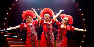 Ons pap wil bij de revue Drie oudere mannen gekleed in lange felrode galajurken en met veren hoofdtooien staan stralend in een showpose.