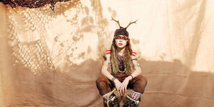 Captain Fantastic Een jongen zit voor een bruin doek. Hij heeft verfstrepen op zijn gezicht en twee stokken op zijn hoofd. Hij ziet er wild uit.