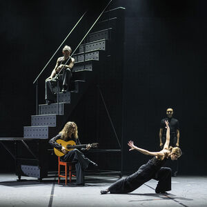 Een danseres staat in snoekduik-pose met armen en vingers als vleugels naar achter gestrekt.
Naast haar zit een gitarist en achter haar drie mannen bij een hoge zwart metalen trap.
