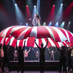 Een acrobate hangt hoog in de lucht boven een grote roodwitte parachute die door vele dansers omhoog wordt gehouden.