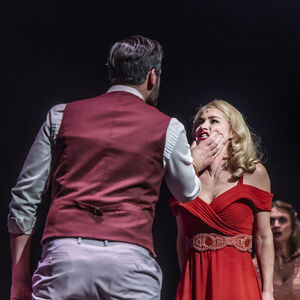 Een man staat met zijn rug naar ons toe. Hij draagt een grijs pak met een rood vest. Hij houdt een vrouw stevig vast in haar gezicht. Hij knijpt in haar wangen. De vrouw kijkt boos en draagt een lange rode jurk. 