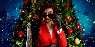 A xxxmas Carola met Nandi van Beurden in een rood pak met zonnebril, staand voor een kerstboom.