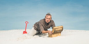 In het zand zit een man op zijn knieën bij een klein kistje met geopende deksel. Achter hem steekt een rood plastic schepje in het zand.