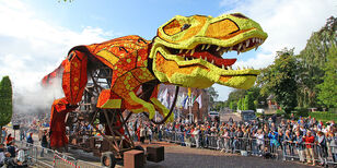 Een hoog boven de bomen uittorende corsowagen van een dino trekt door de straat. De dino is helemaal gemaakt van kleurrijke dahliabloemen.