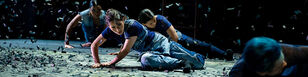 Een drietal dansers liggen op de vloer met confetti om hen heen, het lijkt alsof ze door de wind worden weggeblazen. 