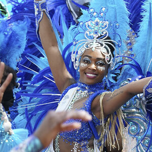 Een vrouw in een blauwe glitter outfit, met veren om haar heen en een kroon. Ze lacht vrolijk in de camera.