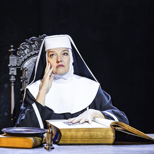 Sanne Wallis de Vries als Moeder Overste in Sister Act. Ze draagt een nonnenhabijt. Ze zit achter een tafel waar een enorm groot boek op ligt. Ze heeft een hand op het boek gelegd, en de andere onder haar hoofd. Ze kijkt bedenkelijk. 