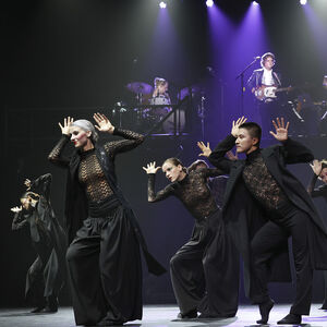 Dansers in zwarte kleding met kanten accenten. Zij dansen met zwaaiende handen. Op de achtergrond de band Blaudzun.