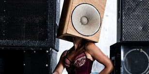 Een vrouw in sexy bordeauxrood ondergoed. Zwarte geluidsboxen om haar heen. Een houten speaker is over haar hoofd geschoven waardoor de speaker haar hoofd lijkt te zijn.