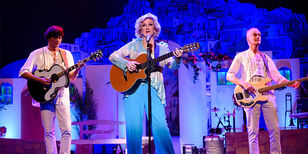 Tineke Schouten speelt gitaar op een blauw toneel. Twee mannen staan naast haar. Ook de twee mannen hebben gitaren vast. Tineke draagt een blauwe blouse. De twee mannen dragen een witte blouse. 