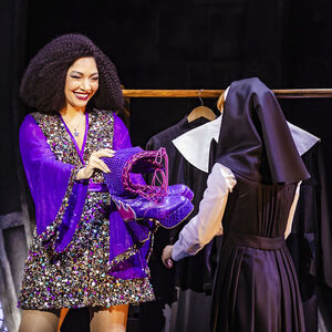 April Darby als Deloris in Sister Act. Deloris draagt een korte glitterjurk met lange, uitlopende, paarse mouwen. Ze houdt een paar torenhoge paarse laarsen vast. Naast haar staat een non. De non staat met haar rug naar de foto.