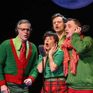 Vier uitbundig zingende mensen rondom een microfoon. Ze dragen groene en rode kleding in kerstsfeer.